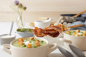 White wooden table with bowls of thick green pea soup embellished with roasted rusks, green peas, slices of grilled bacon, crushed black pepper and cream.