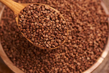 Uncooked buckwheat in a wooden spoon over the bowl with buckwheat. Close up.