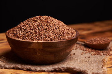 Uncooked buckwheat in a wooden bowl with the wooden spoon full of buckwheat, rustic table. Black background.