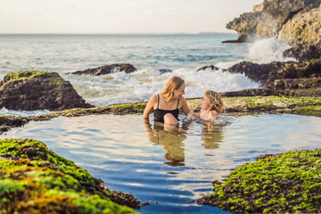 Fototapeta na wymiar Mother and son tourists on Pantai Tegal Wangi Beach sitting in a bath of sea water, Bali Island, Indonesia. Bali Travel Concept. Traveling with children concept. Kids friendly places