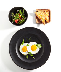 Two fried eggs, cheese toast and green salad mix on black plate top view