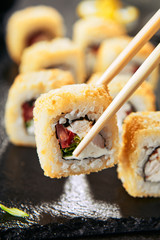 Macro shot of eating tempura uramaki sushi rolls with smoked chicken, cream cheese, green onion, tomato, rice and nori. Chopsticks holding hot crispy maki roll on black stone slate background closeup
