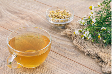 Glass cup of green tea with fresh and dry white chamomile flowers on wooden background.