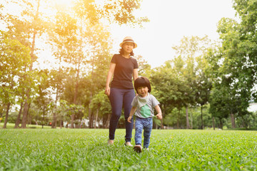 Fototapeta premium Cute 2 year old asian toddler girl exploring the park with excitement while the mother follow and walking behind protective in the natural warm sunlight tone, concept protective mom and child love.