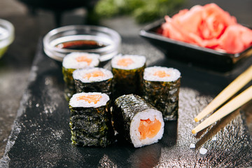 Fototapeta Macro shot of salmon hosomaki sushi on natural black slate plate background with selective focus. Thin maki sushi rolls with raw trout, cucumber, rice, sesame and nori closeup obraz