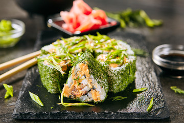 Green uramaki sushi roll with rice, cream cheese, fried salmon, tomatoes, beijing cabbage, green onion, dill and spicy sauce closeup. Macro shot of colorful maki susi rolls on black slate background