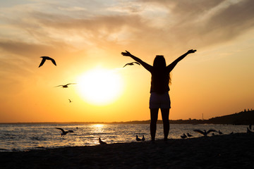 Fototapeta na wymiar A young woman in shorts and a T-shirt is standing at sunset against the background of the sea with her hands up, seagulls are flying around. The concept of summer, sea, relaxation and happiness