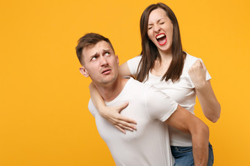 Young couple in white t-shirts posing isolated on yellow orange background. People lifestyle concept. Mock up copy space. Giving piggyback ride to joyful sitting on back, clenching fist like winner.