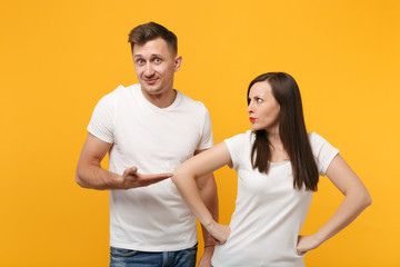 Perplexed young couple two friends guy girl in white empty blank t-shirts posing isolated on yellow orange background. People lifestyle concept. Mock up copy space. Standing with arms akimbo on waist.