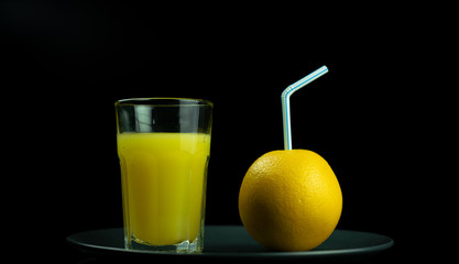 A glass of orange juice stands next to a yellow orange, which is inserted into the tube for drinks. Black background.