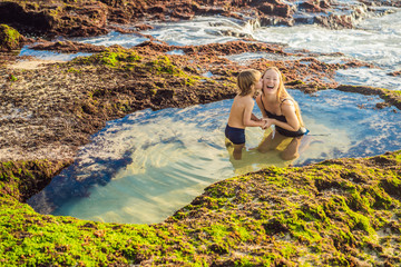 Mother and son tourists on Pantai Tegal Wangi Beach sitting in a bath of sea water, Bali Island, Indonesia. Bali Travel Concept. Traveling with children concept. Kids friendly places