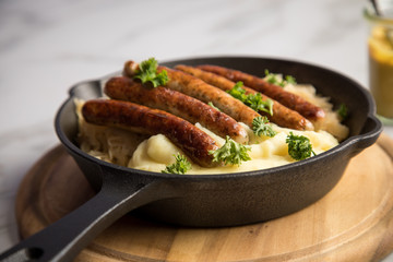 Kleine Bayrische Nürnberger Rostbratwürste mit Sauerkraut, Kartoffelpüree und Senf in rustikaler Gusseisenpfanne auf Holz Brett und Marmor Tisch