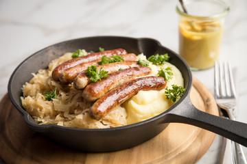 Kleine Bayrische Nürnberger Rostbratwürste mit Sauerkraut, Kartoffelpüree und Senf in rustikaler Gusseisenpfanne auf Holz Brett und Marmor Tisch