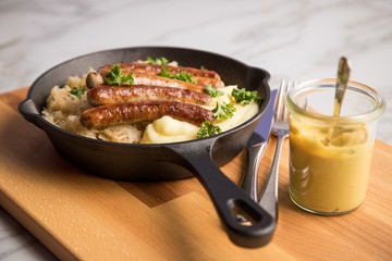 Kleine Bayrische Nürnberger Rostbratwürste mit Sauerkraut, Kartoffelpüree und Senf in rustikaler Gusseisenpfanne auf Holz Brett und Marmor Tisch