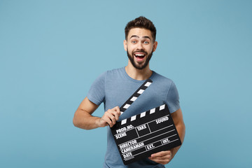 Young surprised man in casual clothes posing isolated on blue wall background, studio portrait. People lifestyle concept. Mock up copy space. Holding in hands classic black film making clapperboard.