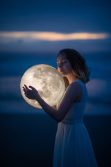 Naklejka premium delikatny obraz anioła, dziewczyny z księżycem w rękach na nocnej plaży. fotografia artystyczna. Bajka i zagadka. Astrologia i znaki zodiaku.