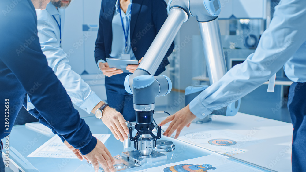 Leinwandbilder Team of Industrial Robotics Engineers Gathered Around Table With Robot Arm, They Use Tablet Computer to Manipulate and Program it to Pick Up and Move Metal Component. Focus on Hands