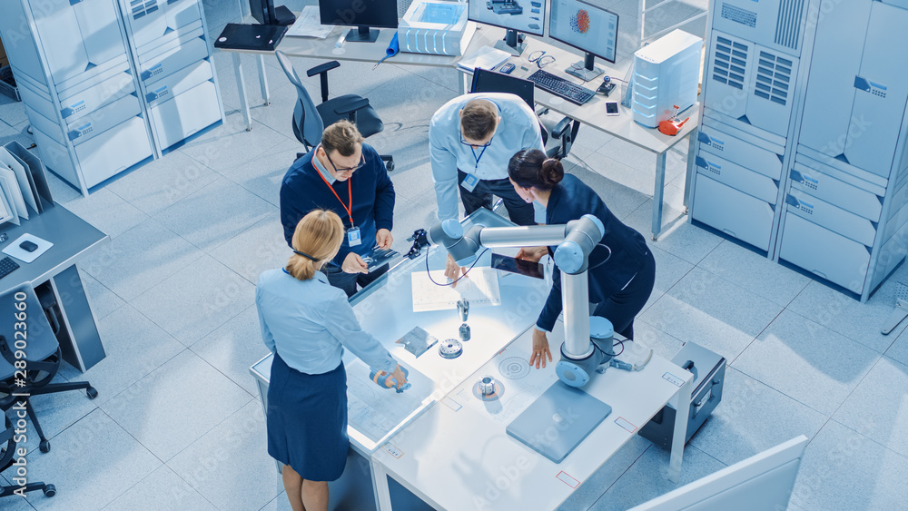 Leinwandbilder Team of Industrial Robotics Engineers Gathered Around Table With Robot Arm, They Use Tablet Computer to Manipulate and Program it to Pick Up and Move Metal Component. Top Down Above Shot