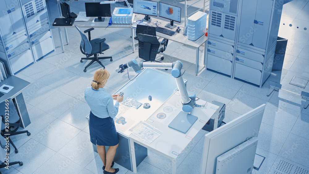 Leinwandbilder Stylish Female Industrial Robotics Engineer Looks at Blueprints Engineering Ways to Program and Manipulate Robot Arm for Lifting, Moving Objects. Technology Research Facility for Cloud Computing
