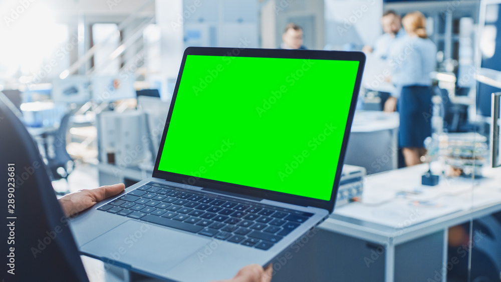 Poster Engineer Holds Green Mock-up Screen Laptop Computer. In the Background Team of Industrial Engineers Working with Functional Robot Arm in Modern Bright Research Facility