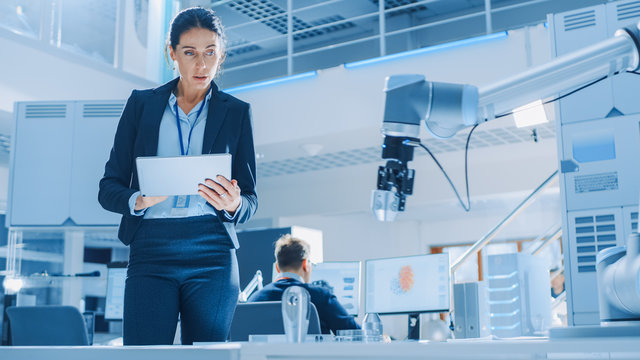 Stylish Female Industrial Robotics Engineer Uses Tablet Computer to Program and Manipulate Robot Arm for Lifting, Moving Objects. Technology Research Facility for Machine Learning and Cloud Computing