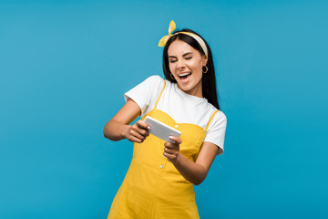 happy woman in yellow dress using smartphone isolated on blue