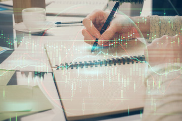 Forex chart hologram on hand taking notes background. Concept of analysis. Double exposure