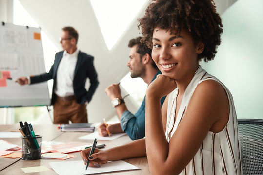 Great working day. Young afro american woman is looking at camera and smiling while her boss standing near whiteboard and discussing something with team