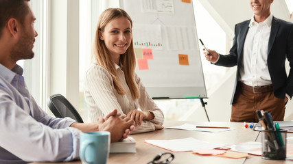 Company progress. Young and cheerful woman looking at camera with smile while her boss standing near whiteboard and explaining something on the background