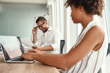 Working together. Young man in formal wear and eyeglasses looking at financial report and discussing it with his afro american female collegue while sitting at the office table