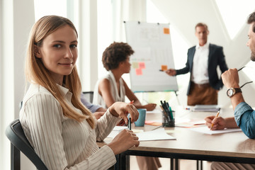 Young woman is looking at camera and smiling while her boss standing near whiteboard and discussing something with team