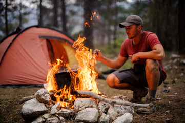 Man in the cap sitting near the pot on the campfire