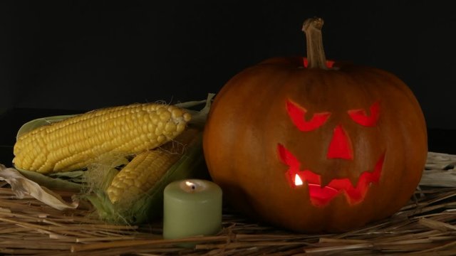 Spooky pumpkin halloween with burning candle and ears of corn lying on the straw, black background