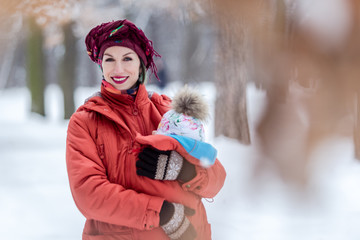 Mother carrying her baby girl wears red jacket and sling.