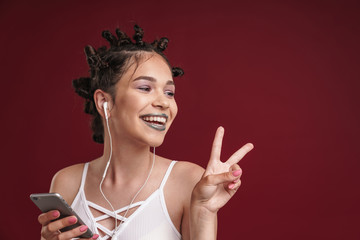 Obraz premium Image of cute punk girl with bizarre hairstyle and dark lipstick showing peace fingers while using smartphone with earphones