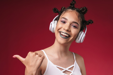 Fototapeta premium Image of joyful punk girl with bizarre hairstyle pointing finger at copyspace while listening to music with headphones