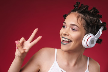 Naklejka premium Image of joyful punk girl with bizarre hairstyle and dark lipstick showing peace fingers while listening to music with headphones