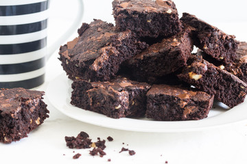Chocolate brownies in a stack on white plate with coffee mug in background on white background - side view