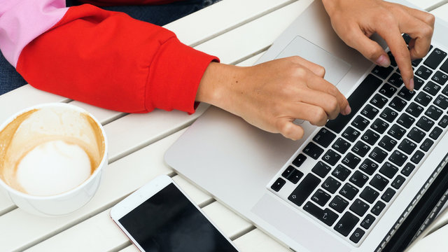 Close Up Of Female Hands Typing On Laptop Keyboard. Working In Cafe. Empty Cup Of Coffee And Mobile Phone Nearby