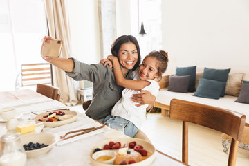 Image of beautiful family mother and little daughter taking selfie photo while having breakfast at home in morning