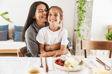 Obraz na płótnie Canvas Image of attractive family mother and little daughter hugging while having breakfast at home in morning