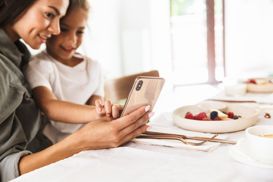 Image of positive family mother and little daughter looking at cellphone while having breakfast at home in morning