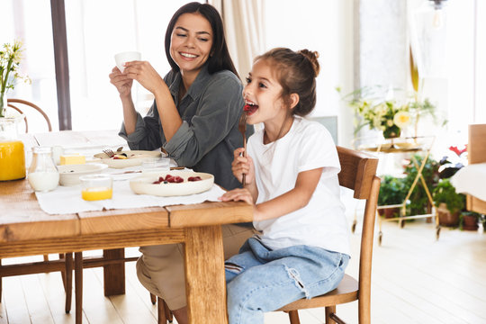 Image of stylish family mother and little daughter eating tasty food together while having breakfast at home in morning