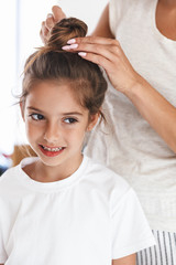 Full length image of beautiful family woman making toptnot hairstyle to her little girl at cozy...