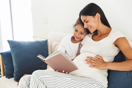 Image of caucasian family pregnant woman and her little daughter reading book while sitting on sofa at home