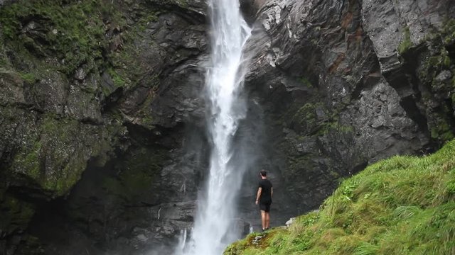 Man standing next to a largewaterfall in the mountains, admiring the power of nature.