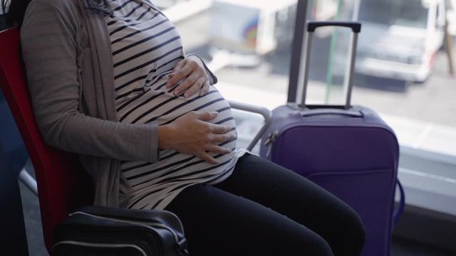 asian pregnant woman sitting beside a suitcase on the airport waiting room before flight departure