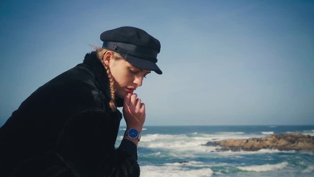 4K UHD Cinemagraph of a young brunette woman sitting by the sea with a black coat and black hat with the waves rushing in the background.