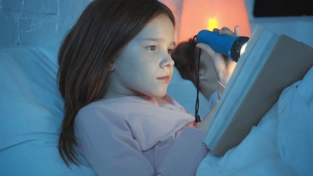daughter reading book with flashlight near sleeping mother