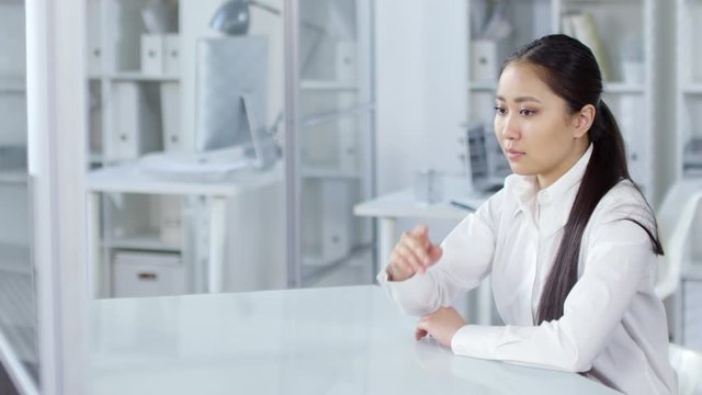 Waist-up shot of young Asian woman in white blouse sitting in office, looking through images on large invisible AR touchscreen with swiping, tapping and enlarging gestures. Suitable for adding AR, VR.